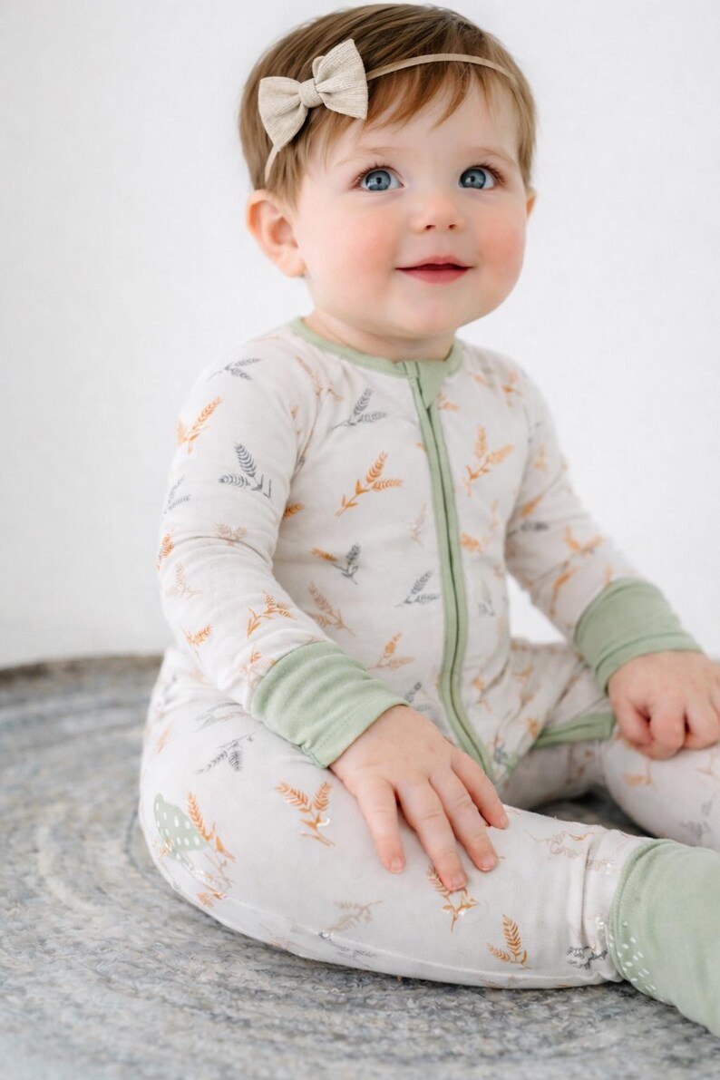 K&ouml;nnte beinhalten: Ein Baby tr&auml;gt einen hellfarbenen Strampler mit gr&uuml;nem Rei&szlig;verschluss und B&uuml;ndchen. Der Strampler hat ein Muster aus Weizen&auml;hren in Orange und Grau. Ein beiges Schleifen-Stirnband ziert den Kopf des Babys.