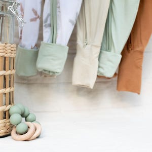 K&ouml;nnte beinhalten: Babykleidung in verschiedenen Farben, darunter Wei&szlig;, Beige und Rost, h&auml;ngt vor einer wei&szlig;en Backsteinmauer. Ein Weidenkorb und ein Glas mit getrockneten Pflanzen befinden sich links. Ein gr&uuml;ner Bei&szlig;ring mit Holzringen ist im Vordergrund.