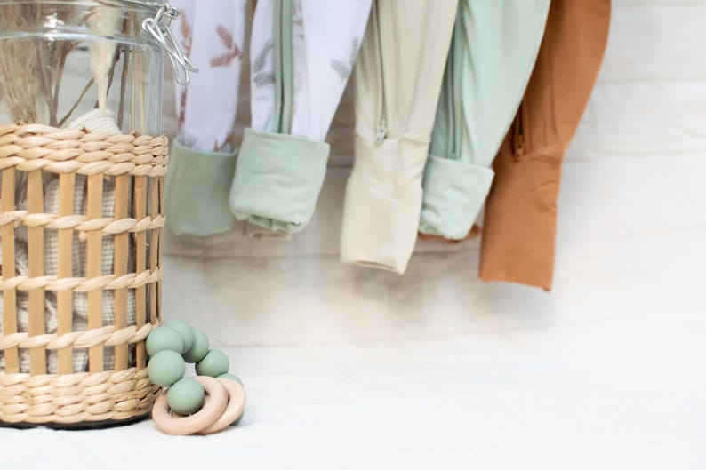 K&ouml;nnte beinhalten: Babykleidung in verschiedenen Farben, darunter Wei&szlig;, Beige und Rost, h&auml;ngt vor einer wei&szlig;en Backsteinmauer. Ein Weidenkorb und ein Glas mit getrockneten Pflanzen befinden sich links. Ein gr&uuml;ner Bei&szlig;ring mit Holzringen ist im Vordergrund.