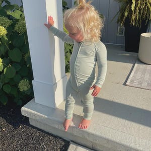 K&ouml;nnte beinhalten: Ein junges Kind in einem hellgr&uuml;nen, lang&auml;rmeligen Overall mit durchgehendem Rei&szlig;verschluss. Das Kind hat blonde Haare und steht drau&szlig;en auf einer Betontreppe. Der Overall bedeckt die Arme und Beine des Kindes.