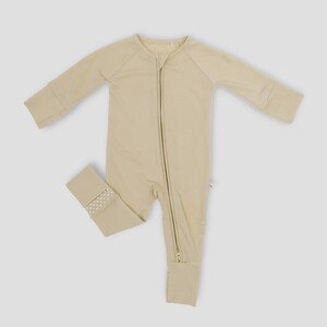K&ouml;nnte beinhalten: Hellbeige Baby-Schlafanzug mit langen &Auml;rmeln und durchgehendem Rei&szlig;verschluss. Der Schlafanzug hat ein schlichtes Design mit gerippten B&uuml;ndchen und einem kleinen gepunkteten Muster am Kn&ouml;chelb&uuml;ndchen. Hergestellt aus einem weichen, bequemen Material.