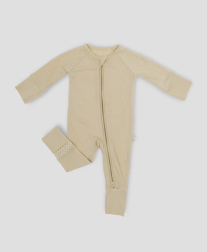 K&ouml;nnte beinhalten: Hellbeige Baby-Schlafanzug mit langen &Auml;rmeln und durchgehendem Rei&szlig;verschluss. Der Schlafanzug hat ein schlichtes Design mit gerippten B&uuml;ndchen und einem kleinen gepunkteten Muster am Kn&ouml;chelb&uuml;ndchen. Hergestellt aus einem weichen, bequemen Material.