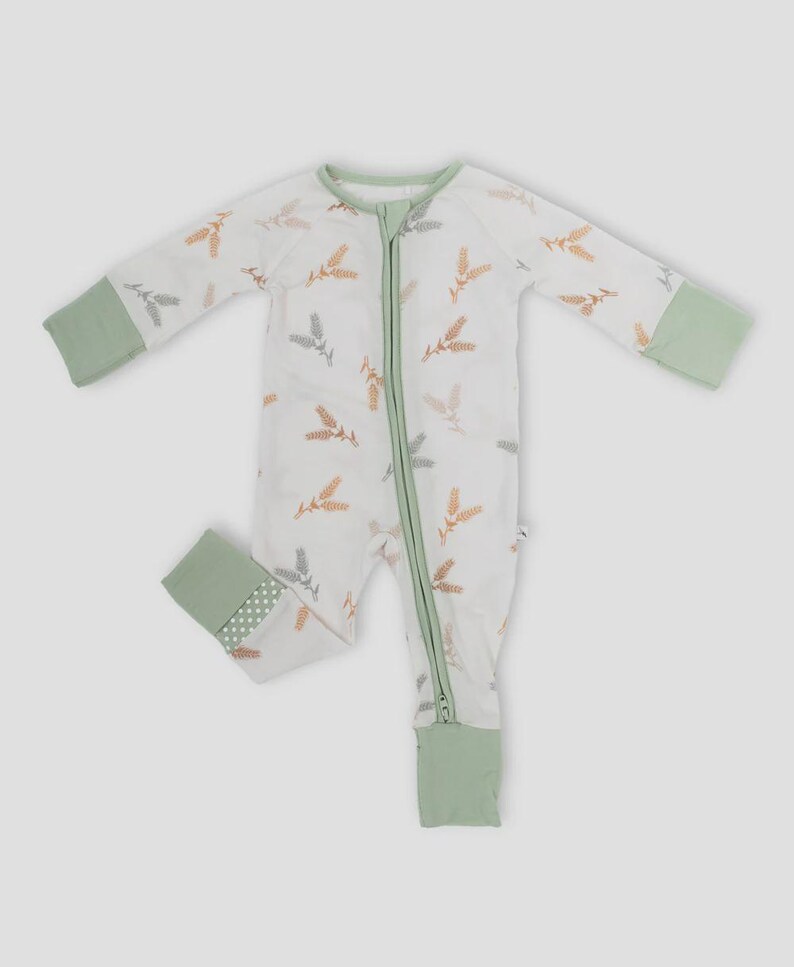 K&ouml;nnte beinhalten: Wei&szlig;er Baby-Schlafanzug mit einem Weizenmuster in Braun-, Grau- und Oranget&ouml;nen. Der Schlafanzug hat einen durchgehenden Rei&szlig;verschluss, gr&uuml;ne Paspeln und gr&uuml;ne B&uuml;ndchen an &Auml;rmeln und F&uuml;&szlig;en. Die Fu&szlig;b&uuml;ndchen haben ein Tupfenmuster.