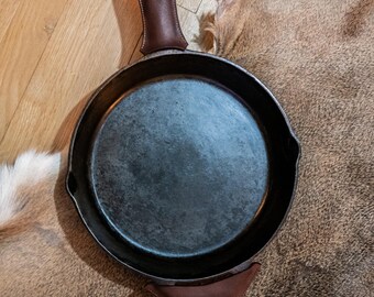 Op de afbeelding: Een ronde zwarte gietijzeren koekenpan met een donkerbruine lederen handgreepbeschermer en een bijpassende bruine lederen zijgreepbeschermer. Het bakoppervlak is glad en donker, met twee kleine schenktuiten aan weerszijden van de panrand.
