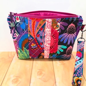 May include: A colorful, quilted cosmetic bag with a vibrant floral and geometric pattern. The bag features a magenta zipper and interior, and a matching wrist strap. The bag is approximately 8 inches wide and 6 inches tall.