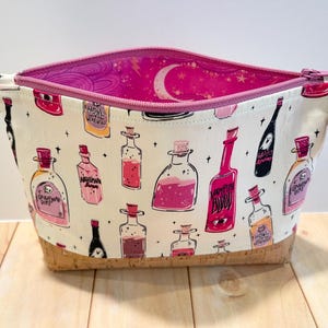 May include: A white cosmetic bag with a cork bottom and a pink interior. The bag is decorated with illustrations of potion bottles in shades of pink, red, and yellow. A silver zipper pull with a pink tassel adds a decorative touch.