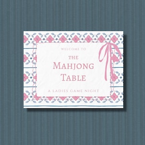 Può includere: Un cartello rettangolare con sfondo bianco e un bordo floreale rosa e blu. Il testo recita: WELCOME TO THE MAHJONG TABLE A LADIES GAME NIGHT. Un'illustrazione di un nastro rosa si trova sul lato destro del bordo, incorniciando il testo centrale.