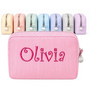 May include: A rectangular pink and white striped zippered pouch featuring the name Olivia embroidered in dark pink script on the front. Seven additional pouches in various pastel colors with matching stripes are arranged in a row behind it.