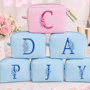 May include: A stack of six rectangular cosmetic bags featuring a pinstripe pattern. One pink bag with a floral letter C and five light blue bags with floral letters D, A, P, J, and Y. A small tag attached to the top bag reads EMBROIDERED.