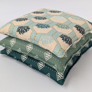 May include: A stack of three square throw pillows. The top pillow features a light blue and beige hexagonal pattern with dark blue floral motifs. The middle pillow is sage green with white geometric diamond shapes. The bottom pillow is dark teal.