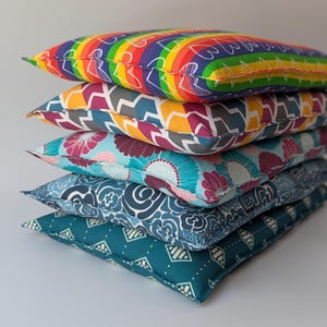 May include: A stack of five decorative pillows with various colorful patterns. The top pillow features a rainbow design, followed by geometric, floral, and paisley prints in shades of blue, pink, and teal. The pillows are rectangular and appear to be made of fabric.