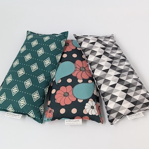 May include: Three rectangular pillows with distinct patterns. The left pillow is teal with white diamond shapes. The center pillow features a dark background with pink flowers, teal paisley shapes, and tan dots. The right pillow has a black, gray, and white triangle pattern.