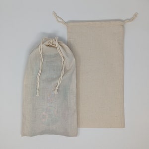 May include: Two off-white drawstring bags. One bag is closed with a drawstring, and the other is open. The bags are made of a natural-colored fabric. The closed bag appears to contain items, while the other is empty.