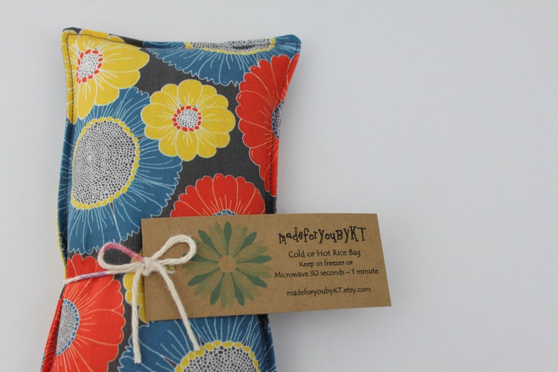 May include: A rectangular, floral-patterned rice bag in shades of blue, yellow, and red, tied with a white string and a brown tag. The tag reads "made for You ByKT" and includes instructions for use. The bag is designed for cold or hot use.