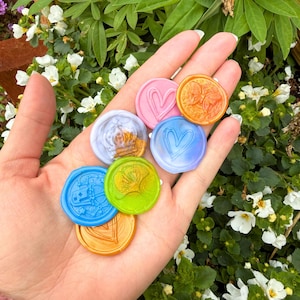May include: A collection of seven circular wax seals in various colors including pink, orange, blue, light purple, and lime green. Each seal features a unique embossed design such as hearts, a ginkgo leaf, and a crown. Text reads: *Colors and designs may vary.