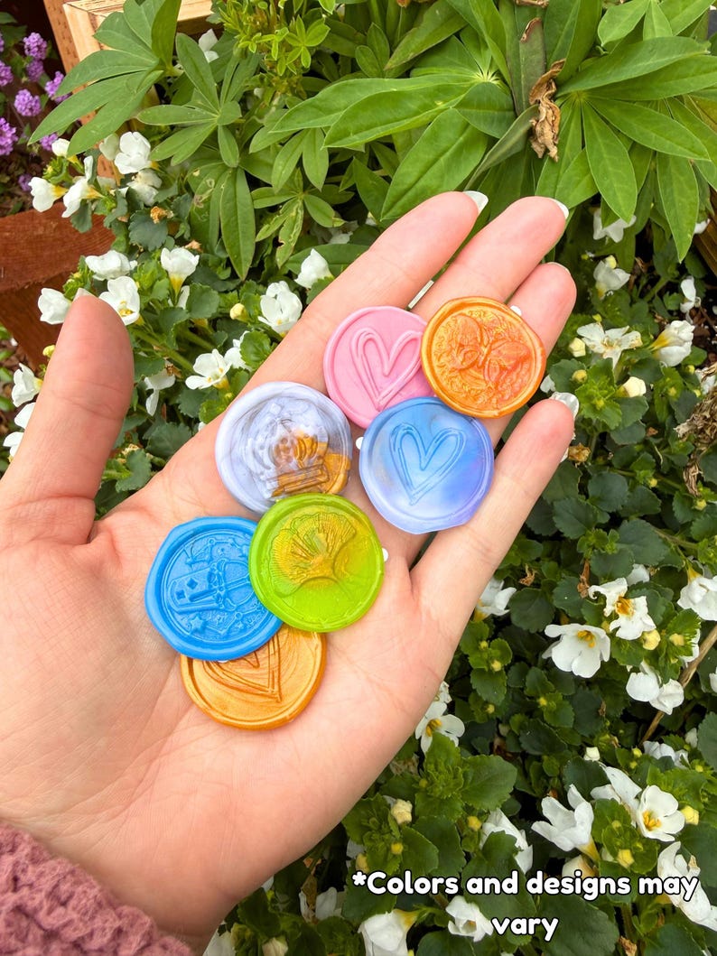 May include: A collection of seven circular wax seals in various colors including pink, orange, blue, light purple, and lime green. Each seal features a unique embossed design such as hearts, a ginkgo leaf, and a crown. Text reads: *Colors and designs may vary.