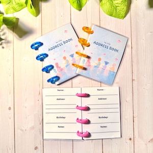 May include: Two small address books with light blue covers featuring a colorful illustration of children and the text "my little ADDRESS BOOK." One book has blue plastic discs, the other orange. An open book with pink discs shows fields for name, address, birthday, and notes.