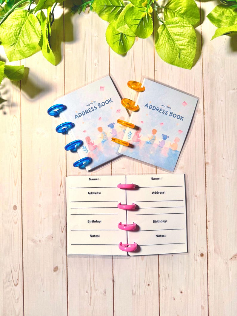 May include: Two small address books with light blue covers featuring a colorful illustration of children and the text "my little ADDRESS BOOK." One book has blue plastic discs, the other orange. An open book with pink discs shows fields for name, address, birthday, and notes.