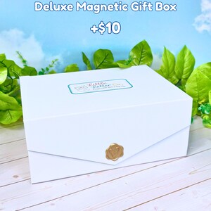 May include: A white rectangular magnetic gift box featuring a gold-colored wax seal on the front flap and a rectangular label on the lid that reads Little Letter Co. a little letter goes a long way. The box is presented against a light blue sky background.