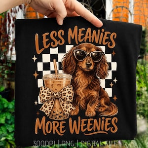 Könnte beinhalten: Ein schwarzes T-Shirt mit einer Grafik eines braunen langhaarigen Dackels mit Sonnenbrille neben einem Bubble Tea mit einer Leopardenmuster-Schleife. Das Design enthält einen schwarz-weißen Schachbrett-Hintergrund, Sternakzente und den Text LESS MEANIES MORE WEENIES.