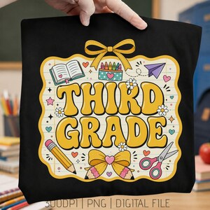 Puede incluir: Un artículo de tela negra con un diseño gráfico que muestra el texto amarillo "THIRD GRADE" dentro de un marco de papel rayado. El diseño incluye ilustraciones de un libro, crayones, un avión de papel, un lápiz, tijeras y lazos amarillos.