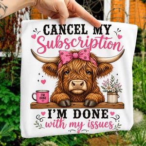 May include: A white t-shirt featuring a brown Highland cow with a pink bow, resting on a wooden plank. The text reads CANCEL MY Subscription, I'M DONE with my issues. A pink mug labeled I'M DONE and a small vase of flowers are on the plank.