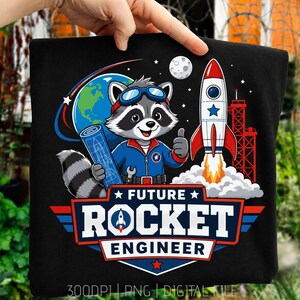 May include: A black t-shirt featuring a graphic of a raccoon dressed as an astronaut with a blue jumpsuit, goggles, and a blueprint. Beside the raccoon is a rocket ship, the Earth, and the moon. The text reads FUTURE ROCKET ENGINEER.