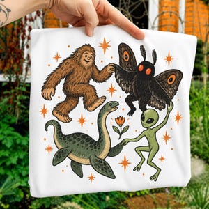Può includere: Una t-shirt bianca con una colorata illustrazione grafica di un Bigfoot marrone, una creatura falena nera, un mostro di Loch Ness verde e un alieno verde che si tengono per mano, circondati da piccole stelle arancioni e un fiore arancione.