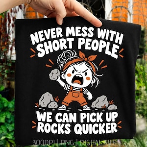 May include: A black t-shirt featuring a graphic of an angry cartoon character with a messy bun and orange overalls holding a rock. The text reads: NEVER MESS WITH SHORT PEOPLE WE CAN PICK UP ROCKS QUICKER. The design includes rocks and orange accents.