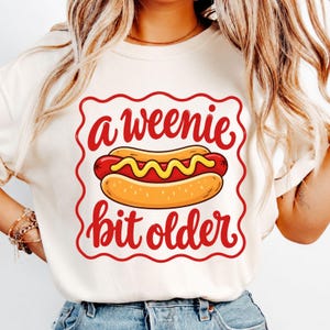 Puede incluir: Una camiseta de color crema con un marco cuadrado festoneado rojo que contiene un gráfico de un perrito caliente con mostaza amarilla. El texto "a weenie bit older" está impreso en letras cursivas rojas sobre y bajo el perrito caliente.