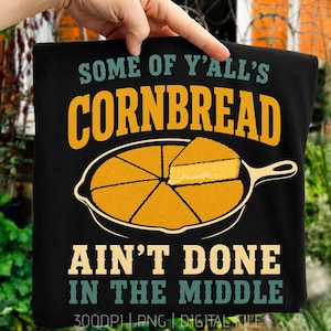May include: A black t-shirt featuring a graphic of a cast iron skillet filled with sliced yellow cornbread. The text reads "SOME OF Y'ALL'S CORNBREAD AIN'T DONE IN THE MIDDLE" in bold yellow and teal lettering, centered above and below the skillet illustration.