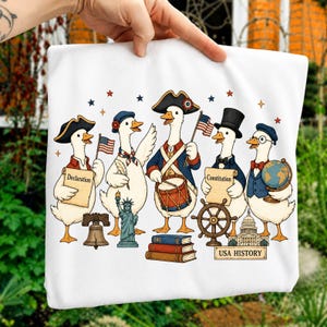 Op de afbeelding: Een wit t-shirt met een grafische illustratie van vijf ganzen in historische Amerikaanse kleding. De ganzen houden voorwerpen vast, waaronder een document met de Declaration, een Constitution-document, een trommel, een vlag en een wereldbol. Iconen zijn de Liberty Bell, het Vrijheidsbeeld, een scheepsroer, het Capitool en de tekst USA HISTORY.
