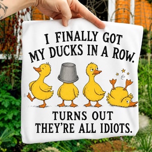 May include: A white t-shirt featuring a graphic of four yellow ducks and the black text "I FINALLY GOT MY DUCKS IN A ROW. TURNS OUT THEY'RE ALL IDIOTS." The ducks are depicted in various states: one walking, one with a bucket on its head, one standing, and one upside down.