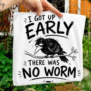 May include: A white t-shirt featuring a black graphic illustration of a tired-looking crow perched on a branch. The text above the crow reads I GOT UP EARLY and the text below the crow reads THERE WAS NO WORM in a bold, hand-drawn style font.