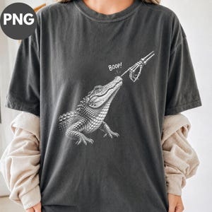 Może przedstawiać: Ciemnoszary t-shirt z krótkim rękawem z białą grafiką przedstawiającą szkieletową dłoń dotykającą nosa aligatora. Napis "Boop!" jest nadrukowany na biało nad pyskiem aligatora. Koszulka posiada okrągły dekolt.