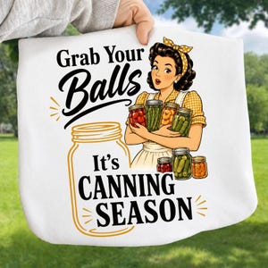 Pode incluir: Uma t-shirt branca com um gráfico de uma pessoa de cabelo escuro e fita amarela segurando vários frascos de vidro com vegetais em conserva. O texto diz Grab Your Balls It's CANNING SEASON em letras pretas e amarelas.
