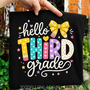 Pode incluir: Uma t-shirt preta com um gráfico colorido com o texto hello THIRD grade. O design inclui um laço amarelo em forma de lápis, letras com bolinhas em amarelo, turquesa, rosa e roxo, e pequenos detalhes de estrelas e corações.