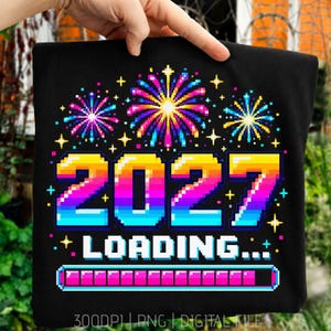 May include: A black t-shirt featuring a pixel art graphic of colorful fireworks above the text 2027 LOADING... and a progress bar. The design uses a vibrant gradient of yellow, orange, pink, purple, and blue colors against the dark fabric.