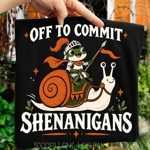 Op de afbeelding: Een zwart t-shirt met een afbeelding van een groene kikker in een ridderhelm en cape, rijdend op een grote slak. De kikker houdt een speer vast. De tekst OFF TO COMMIT SHENANIGANS staat in witte hoofdletters op het shirt gedrukt.