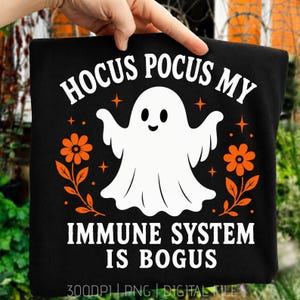 May include: A black t-shirt featuring a white ghost graphic in the center, flanked by orange flowers and stars. The text reads HOCUS POCUS MY IMMUNE SYSTEM IS BOGUS in white capital letters. The design includes small orange stars and floral elements.