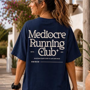 May include: A navy blue short-sleeve t-shirt featuring white text on the back that reads Mediocre Running Club, ESTD. 1991, and ENJOYING EVERY STEP AT OUR OWN PACE, accompanied by small star icons and a horizontal line with five stars below the text.