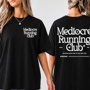 May include: A black t-shirt featuring white text. The front left chest displays the words Mediocre Running Club with small star icons. The back features the same text in a larger font, with ESTD. 1991, the phrase ENJOYING EVERY STEP AT OUR OWN PACE, and five stars.