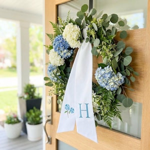 May include: A floral wreath featuring white and blue hydrangeas, eucalyptus, and fern foliage. A white ribbon with the letter "H" embroidered in blue is attached. The wreath is displayed on a wooden door with a glass panel.