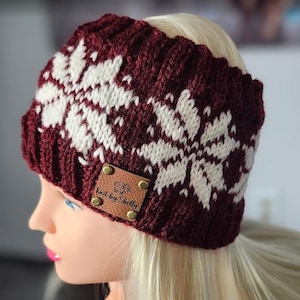 May include: A burgundy and white knit headband with a snowflake design. The headband features a ribbed texture and a small brown leather tag with the text "Knit by Shelly". This winter accessory is designed to keep the ears warm.