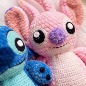 May include: Close-up of two handmade crochet plush toys. One is pink with a purple nose and large black eyes, the other is blue. Both have a soft, textured appearance, resembling characters from a popular animated film.