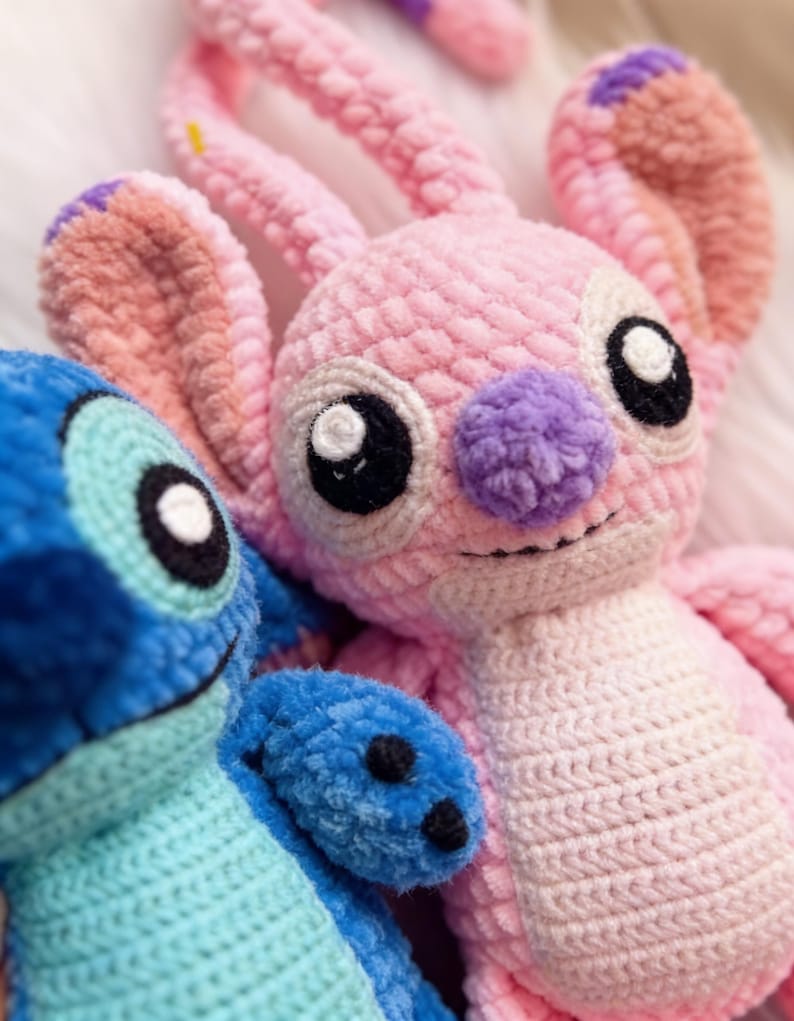 May include: Close-up of two handmade crochet plush toys. One is pink with a purple nose and large black eyes, the other is blue. Both have a soft, textured appearance, resembling characters from a popular animated film.