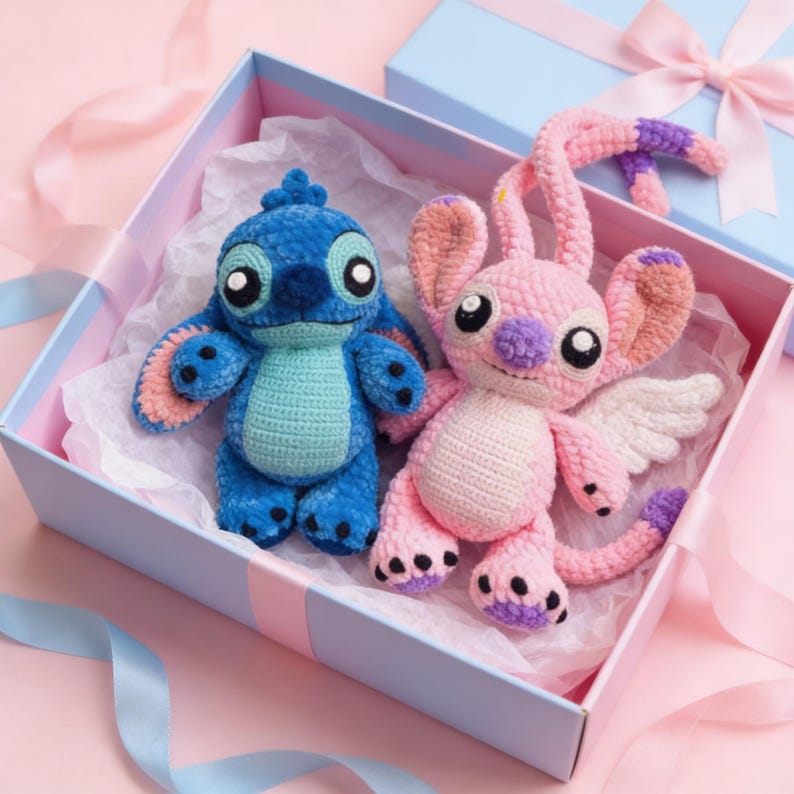 May include: Two handmade crochet plush toys, one blue and one pink, resembling cartoon characters, are presented in a light blue gift box with a pink ribbon. The blue toy has large black eyes and a turquoise belly. The pink toy has white wings.