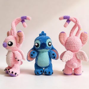 May include: Three handmade crochet figures: two pink creatures with large ears and purple accents, and a blue creature with large eyes. The figures are standing upright, showcasing their detailed craftsmanship and vibrant colors. The toys are soft and plush.