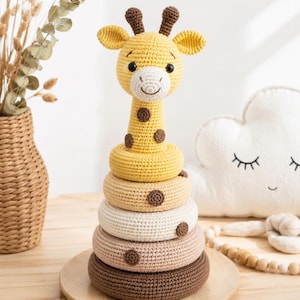 May include: A handmade crochet stacking toy shaped like a giraffe. The giraffe has a yellow head and neck with brown spots and ears. The stacking rings are in shades of yellow, beige, and brown. The toy sits on a wooden base.