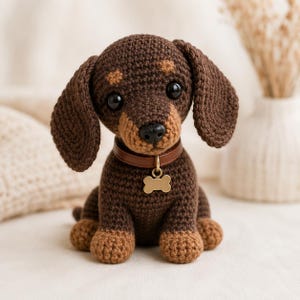 May include: A crocheted dachshund dog in brown and tan hues. The dog features large, floppy ears, a brown leather collar, and a gold-coloured bone-shaped tag. The dog is sitting upright with its paws forward, and its eyes are black.
